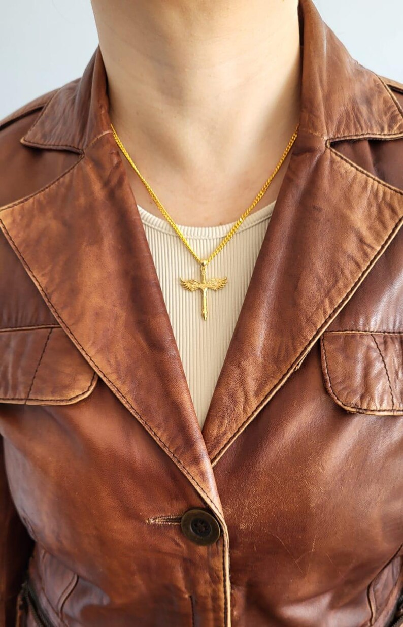 May include: A gold-toned necklace featuring a pendant shaped like a sword with outstretched wings. The pendant hangs from a delicate gold-toned chain, resting against a light-colored ribbed top worn underneath a brown leather jacket with a button.