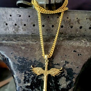 May include: A gold-toned necklace featuring a pendant shaped like a sword with outstretched wings. The pendant is suspended from a gold-toned curb chain. The sword design includes detailed feather textures on the wings and a central hilt.