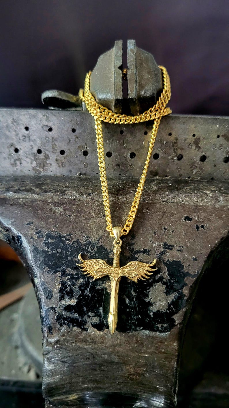 May include: A gold-toned necklace featuring a pendant shaped like a sword with outstretched wings. The pendant is suspended from a gold-toned curb chain. The sword design includes detailed feather textures on the wings and a central hilt.