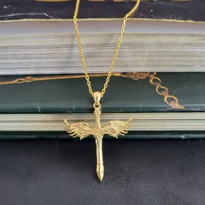 May include: A gold-toned necklace featuring a pendant shaped like a sword with detailed wings extending from the hilt. The pendant hangs from a delicate gold-toned chain. The sword blade is pointed downward, and the wings are textured with feathers.