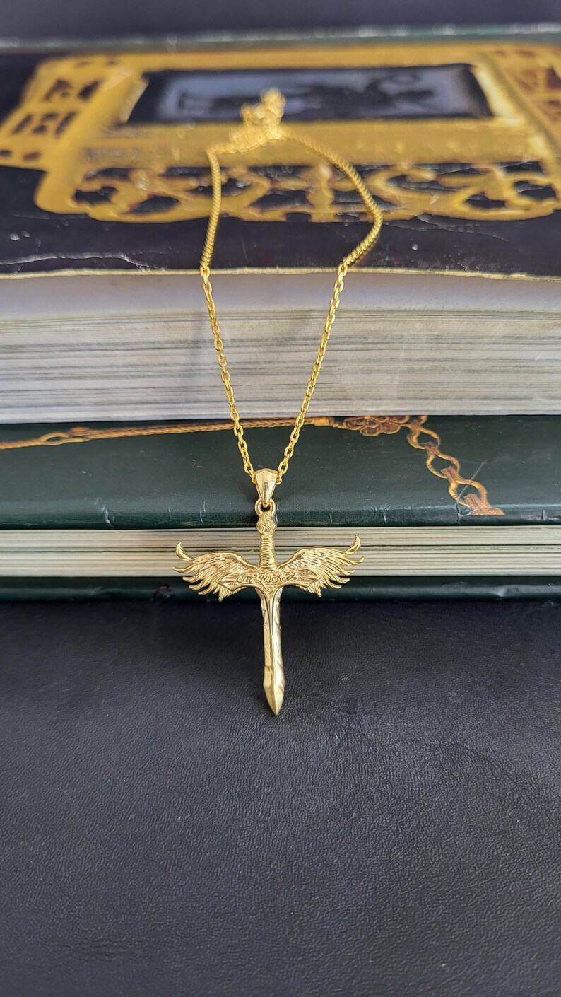May include: A gold-toned necklace featuring a pendant shaped like a sword with detailed wings extending from the hilt. The pendant hangs from a delicate gold-toned chain. The sword blade is pointed downward, and the wings are textured with feathers.