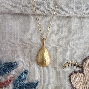 May include: A gold-toned necklace featuring a small, teardrop-shaped pendant with a hammered, textured surface. The pendant hangs from a delicate, thin metal chain. The teardrop shape is rounded at the base and tapers to a point at the top.