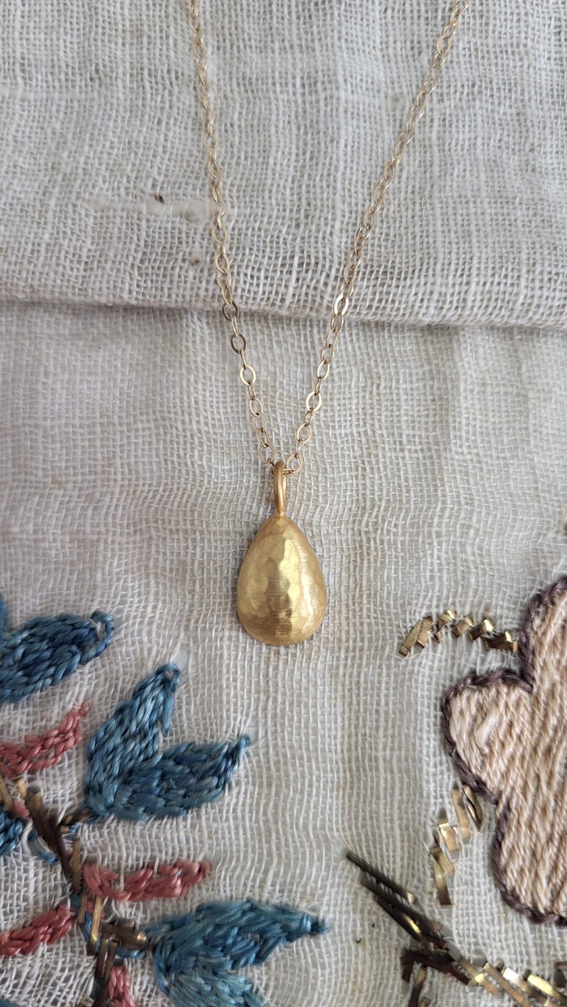 May include: A gold-toned necklace featuring a small, teardrop-shaped pendant with a hammered, textured surface. The pendant hangs from a delicate, thin metal chain. The teardrop shape is rounded at the base and tapers to a point at the top.