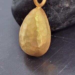 May include: A teardrop-shaped gold pendant featuring a textured, hammered surface finish. The pendant includes a small, circular gold jump ring at the top for attachment, showcasing a matte, brushed appearance across its entire exterior surface.