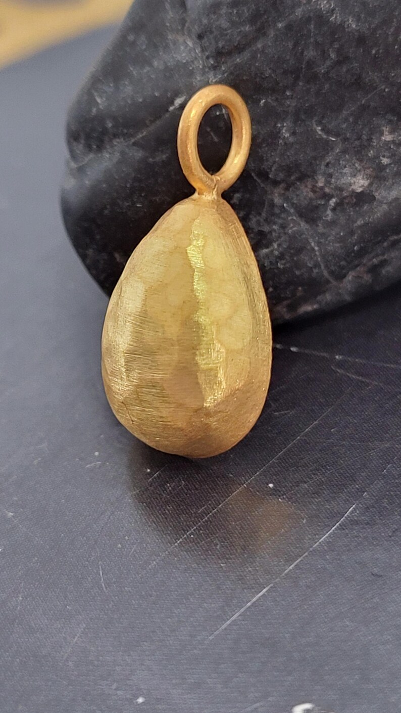 May include: A teardrop-shaped gold pendant featuring a textured, hammered surface finish. The pendant includes a small, circular gold jump ring at the top for attachment, showcasing a matte, brushed appearance across its entire exterior surface.