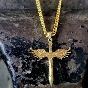 May include: A gold-colored metal necklace featuring a pendant shaped like a sword with detailed wings extending from the hilt. The pendant hangs from a gold-colored curb chain. The sword design includes intricate engravings on the wings and blade.