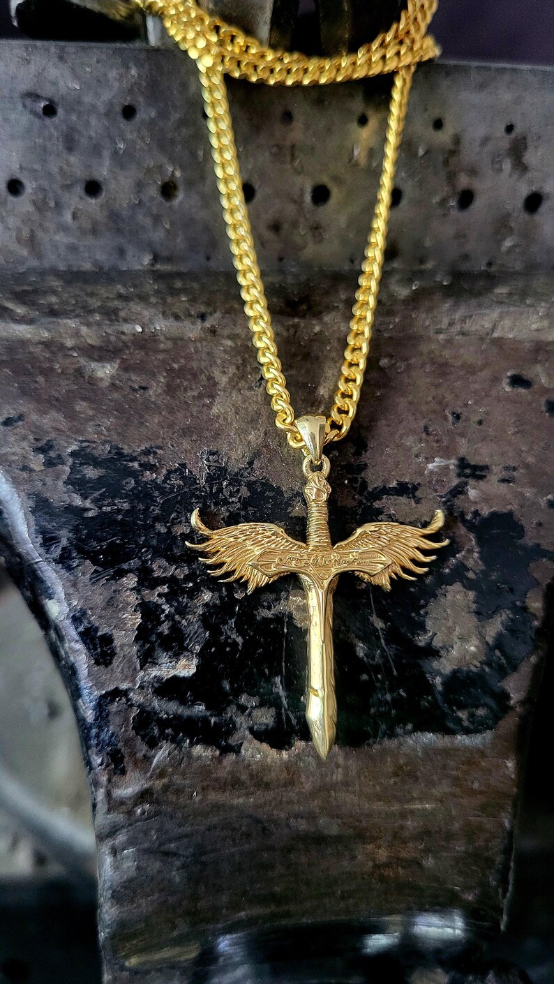 May include: A gold-colored metal necklace featuring a pendant shaped like a sword with detailed wings extending from the hilt. The pendant hangs from a gold-colored curb chain. The sword design includes intricate engravings on the wings and blade.