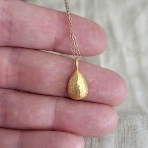 May include: A gold-toned necklace featuring a small, teardrop-shaped pendant with a textured, hammered surface. The pendant hangs from a delicate, thin metal chain. The design is simple and focuses on the metallic finish of the teardrop charm.