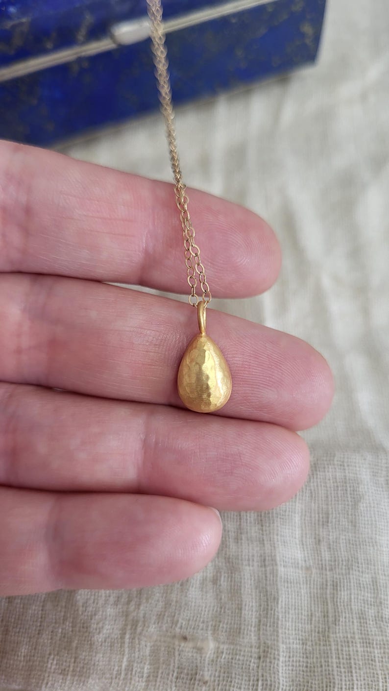 May include: A gold-toned necklace featuring a small, teardrop-shaped pendant with a textured, hammered surface. The pendant hangs from a delicate, thin metal chain. The design is simple and focuses on the metallic finish of the teardrop charm.