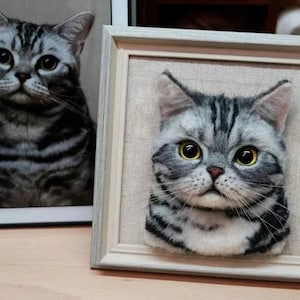May include: A framed needle felted portrait of a grey and white tabby cat. The cat has green eyes and a serious expression.