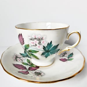 May include: A white teacup and saucer set with a floral design. The cup and saucer have a gold trim and feature pink and green flowers with purple leaves.