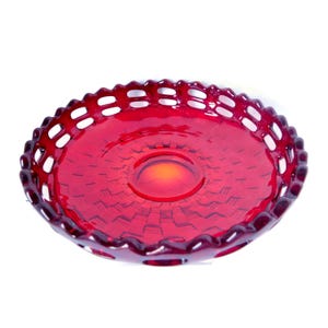 May include: A ruby red glass serving dish with a decorative, openwork rim. The dish has a circular shape with a slightly raised center and a textured pattern on the interior. The color is a deep, transparent red.