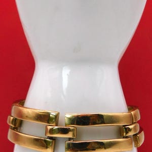 May include: A gold-toned bracelet with a rectangular link design. The bracelet is displayed on a white mannequin arm against a red background. The links are connected in a horizontal pattern.