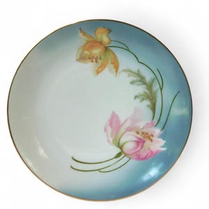 May include: A round, white porcelain plate with a gold rim. The plate features a hand-painted floral design with orange and pink flowers, green leaves, and a light blue background.