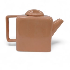 Peut inclure: Une th&eacute;i&egrave;re en c&eacute;ramique de couleur p&ecirc;che avec un corps carr&eacute; et un bec verseur. La th&eacute;i&egrave;re a un design simple et moderne.