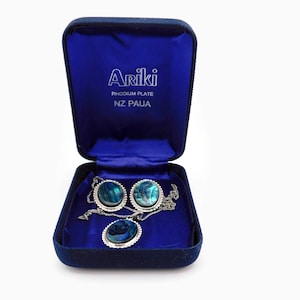 May include: A blue velvet jewelry box with a silver necklace and earrings featuring abalone shell inlays. The box has the text "Ariki Rhodium Plate NZ Paua" on the lid.