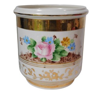 May include: A white ceramic planter with a gold rim and floral design. The planter features a pink rose, blue and yellow flowers, and green leaves. Gold accents and a decorative pattern adorn the base.