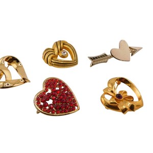 May include: A collection of heart-shaped brooches in gold and red. The brooches include a gold heart with a clear gem, a heart pierced by an arrow, and a heart covered in red rhinestones. These are perfect for Valentine's Day.