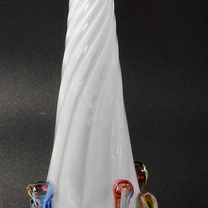 May include: A tall, white glass vase with a spiraled design on the main body. The base features colorful, curved glass accents in shades of blue, yellow, red, and brown. The vase stands on a clear, circular base.