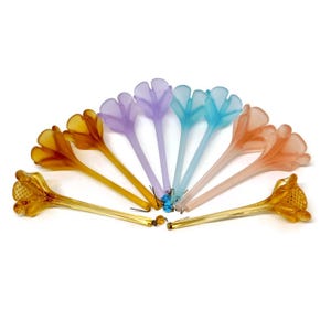 May include: A collection of colorful, flower-shaped glass stir sticks. The colors include shades of yellow, purple, blue, and orange. Each stick has a long stem and a detailed flower head. The sticks are arranged in a fan shape.