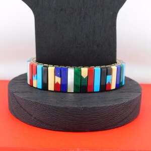 May include: A colorful bracelet with rectangular segments in various colors, including red, blue, black, and green. The bracelet is displayed on a black wooden stand, with a red surface in the background. The bracelet appears to be made of stone or a similar material.