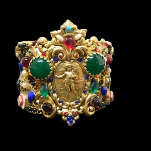 May include: A wide gold-tone cuff bracelet featuring an ornate central oval medallion with a relief of a cherub. The surface is decorated with large round green cabochons, smaller red, blue, and black stones, and white pearl-like accents.