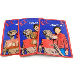 May include: Three unopened packages of E.T. Jewelry. Each package features a blue background with a picture of E.T. and a young boy. The packages are labeled "E.T. Jewelry" and "Star Power, A Division of A&W Ent. Inc."