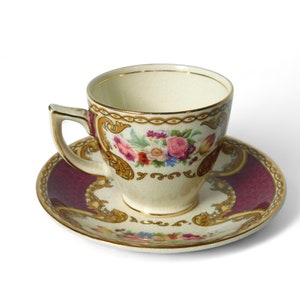 May include: A white teacup and saucer with a floral pattern in shades of pink, yellow, and green. The saucer has a burgundy background with gold accents.