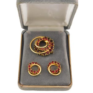 May include: A set of jewelry in a gray velvet box. The set includes a brooch and a pair of earrings. The jewelry is made of gold-toned metal and features red rhinestones.