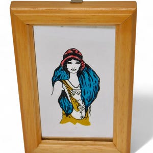 May include: A framed illustration of a woman wearing a red and white hat and a blue scarf. The woman is holding a glass of wine in her hand. The illustration is on a white background and is framed in a light wood frame.