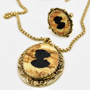 May include: A gold chain necklace with a pendant featuring a cameo of a woman's silhouette. The cameo is set in a gold oval frame with intricate details. The necklace also includes a matching ring with a cameo of a woman's silhouette.
