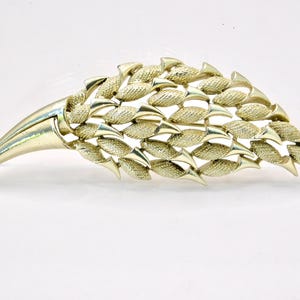 May include: A gold-toned, stylized brooch with a textured, layered design. The brooch has a curved, pointed shape, with a series of textured, leaf-like elements. The overall design is reminiscent of a feather or a stylized plant form.