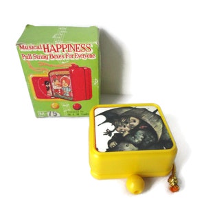 May include: Yellow pull string music box with a picture of two children under an umbrella. The box says "Musical Happiness Pull String Boxes For Everyone" on the box.