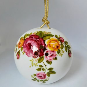 May include: White ceramic ornament with a gold string for hanging. The ornament is decorated with a floral pattern of pink, red, and yellow roses with green leaves.