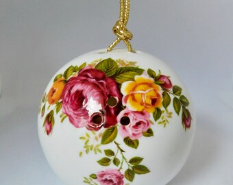 May include: White ceramic ornament with a gold string for hanging. The ornament is decorated with a floral pattern of pink, red, and yellow roses with green leaves.