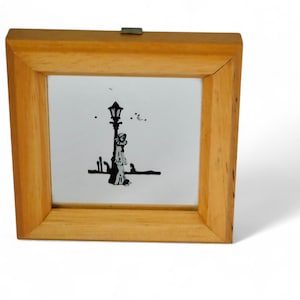 May include: A framed black and white illustration of a person standing under a street lamp. The frame is made of light wood.