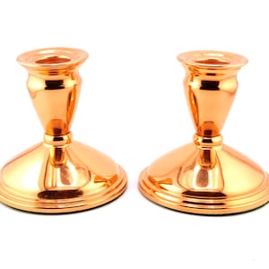 May include: A pair of matching candle holders with a polished copper finish. Each holder features a wide, circular base that tapers upward into a slender neck, topped with a flared rim designed to securely hold a standard taper candle.