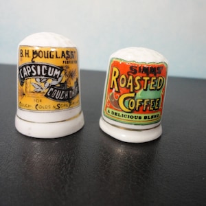 May include: Two vintage ceramic thimbles. One is white with a yellow, orange, and black label that reads "B.H. DOUGLASS CAPSICUM FOR COUCH DROPS COUGH COLDS & SORE THROAT PERFECTLY HARMLESS". The other is white with a yellow, orange, and green label that reads "SIMMS ROASTED COFFEE A DELICIOUS BLEND".
