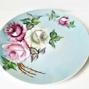 May include: A vintage porcelain plate with a hand-painted design of pink and white roses with green leaves on a light blue background.