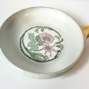 May include: A white porcelain dish with gold handles and a floral design. The design features a pink flower with green leaves and stems.