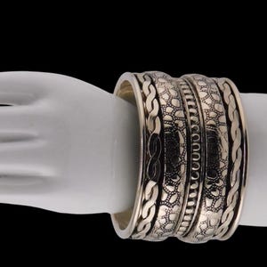 May include: A set of silver-colored bangles with intricate patterns. The bangles feature a combination of braided and textured designs. The jewelry is displayed on a white surface against a black background, highlighting the metallic shine.