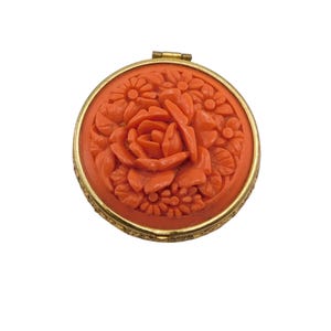 May include: A round gold-tone metal compact mirror featuring a vibrant orange, carved floral relief on the lid. The center of the lid displays a prominent rose surrounded by smaller daisy-like flowers and leaves, all set within a metallic frame.
