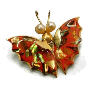 May include: A gold-tone butterfly brooch with red and iridescent wings. The wings are made of abalone shell and have a textured surface. The butterfly has two pearl accents on its head.