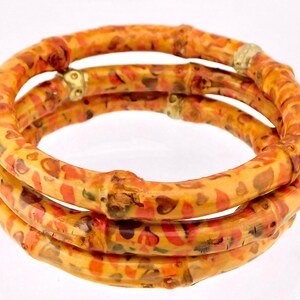 May include: A set of three stacked bangles featuring a bamboo-like texture with a yellow, orange, and brown mottled pattern. Each bangle is accented with small, metallic gold-toned beads integrated into the circular structure of the jewelry.