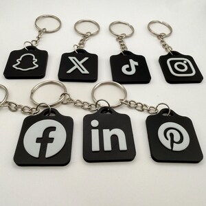 Könnte beinhalten: Eine Sammlung von sieben schwarzen, quadratischen Schlüsselanhängern mit weißen, erhabenen Social-Media-Symbolen für Snapchat, X, TikTok, Instagram, Facebook, LinkedIn und Pinterest. Jeder Anhänger hat einen silbernen Metallring.