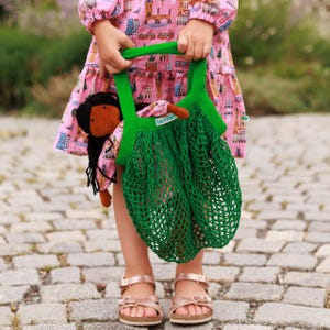 May include: A child is holding a green mesh bag with a doll inside. The bag has green handles and the doll has brown skin and black hair.