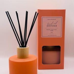 May include: An orange reed oil diffuser with black reeds and a wooden collar. The product is packaged in a matching orange box with the text "Aurora Botanicals Reed Oil Diffuser" and the scent "Rhubarb + Pink Orchid + Vanilla."