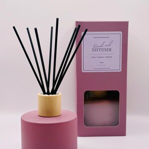 May include: A mauve reed oil diffuser with black reeds and a wooden collar. The diffuser is cylindrical and sits next to a matching box with a window. The box label reads "Reed oil DIFFUSER" with the scent notes "Plum • Jasmine • Almond".
