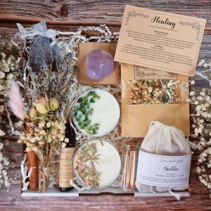 May include: A wooden box filled with self-care items. Includes candles, a purple crystal, dried flowers, a small bottle, and a bag with the word "Goddess" on it. A card with the word "Healing" is also included.