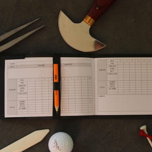 May include: A golf scorebook with an orange pencil, golf ball, tees, and golf club. The scorebook is open, displaying a grid for recording scores. Other tools, including a knife and a metal tool, are also present.